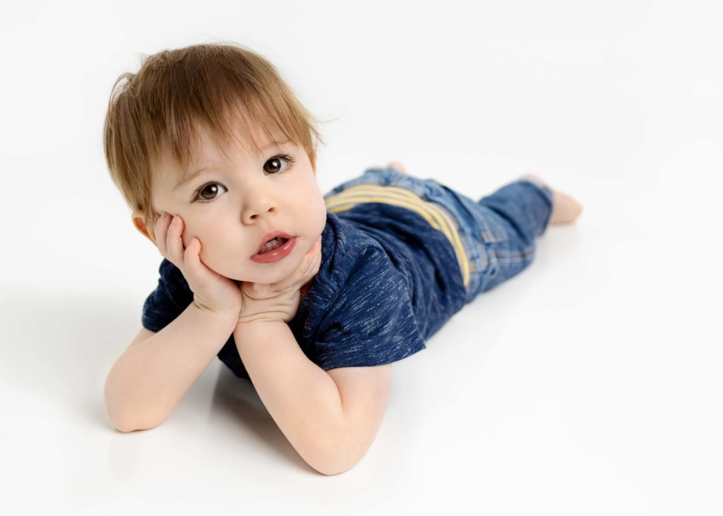 Little boy on his tummy in a navy t-shirt and jeans with his head in his hands looking at the camera by a Flower Mound Photographer