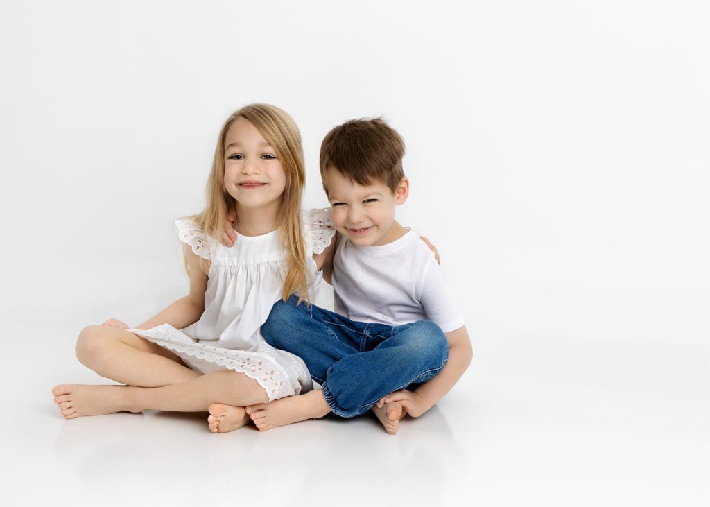 Little girl in white dress sitting with an arm around her little brother in white t-shirt and jeans, smiling at the camera.