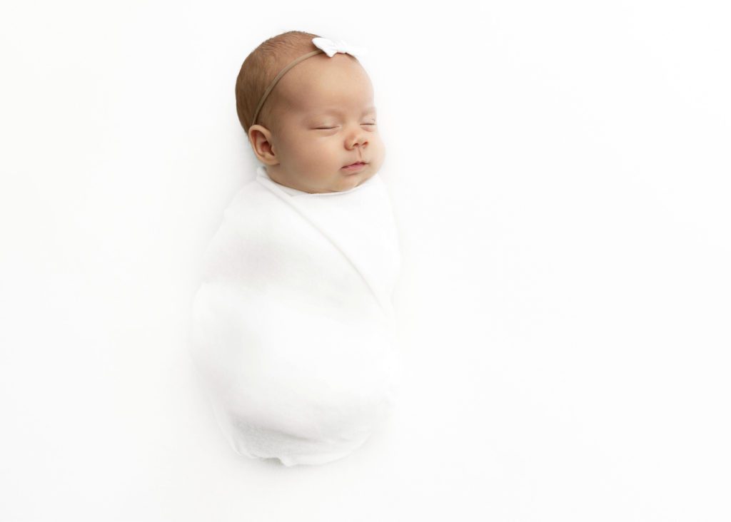 Newborn baby girl wrapped in white cloth with a white bow headband by Fort Worth Newborn Photographer