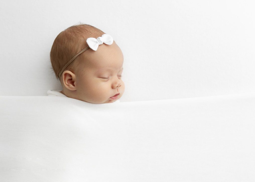 Newborn girl with a white bow headband sleeping under a white blanket with her head turned by Flower Mound photographer.