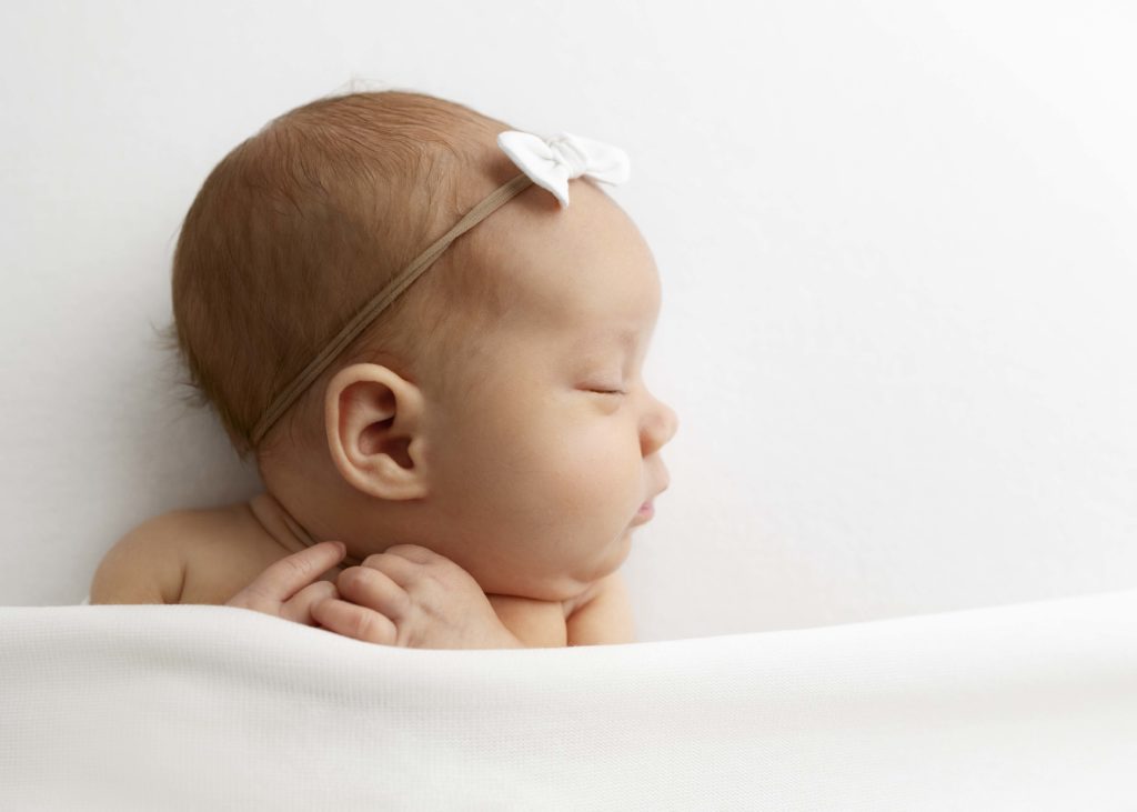 Newborn girl sleeping under a white blanket with a white bow in her hair and head turned.