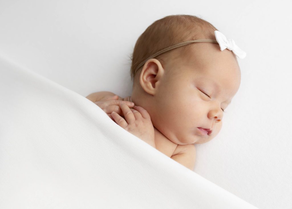 Newborn girl on an angle with hands close to her face and a white bow headband by Flower Mound photographer.