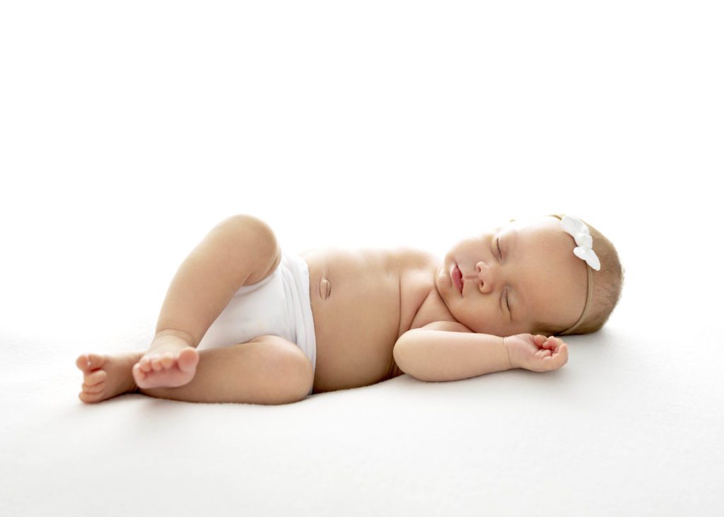 Newborn girl in white diaper and white headband bow sleeping on back with backlighting by 