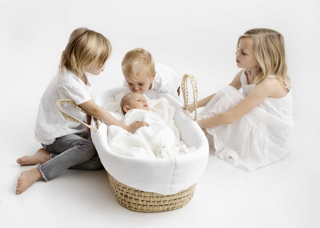 Newborn baby wrapped in white blankets in Moses basket surrounded by three siblings dressed in white