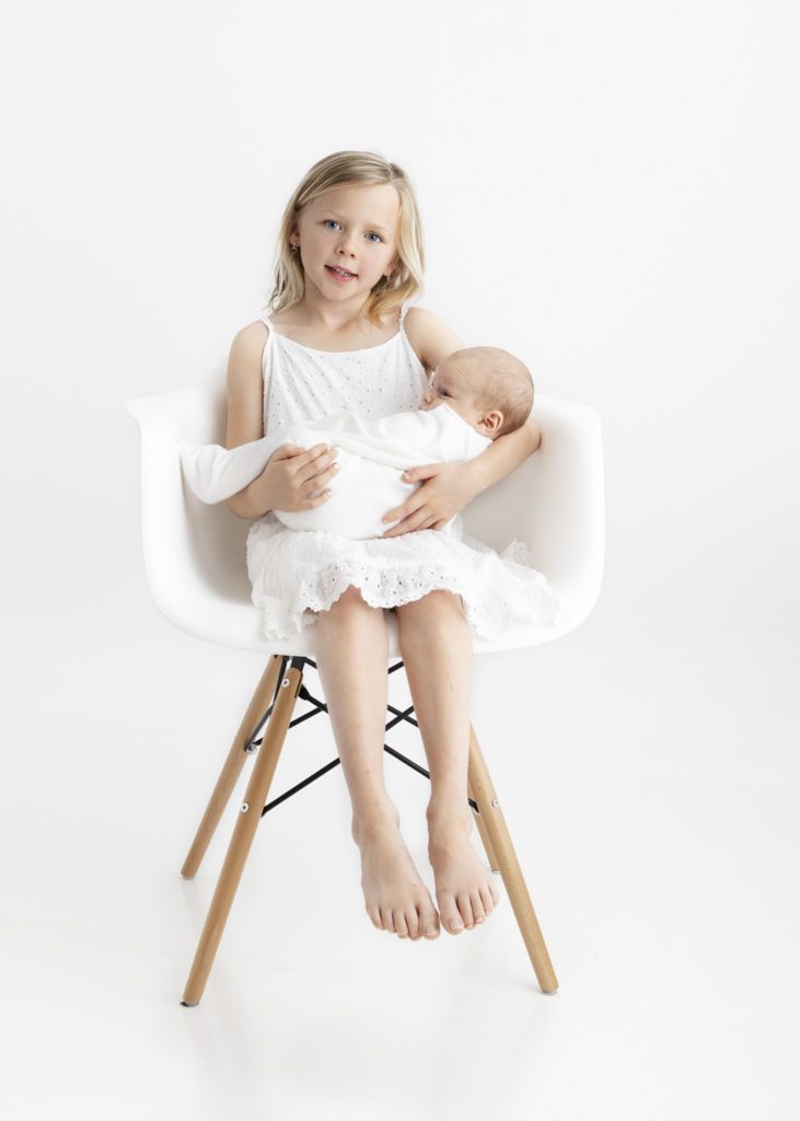 Little girl dressed in white holding her newborn baby brother while sitting in white chair by Grapevine Newborn Photographer