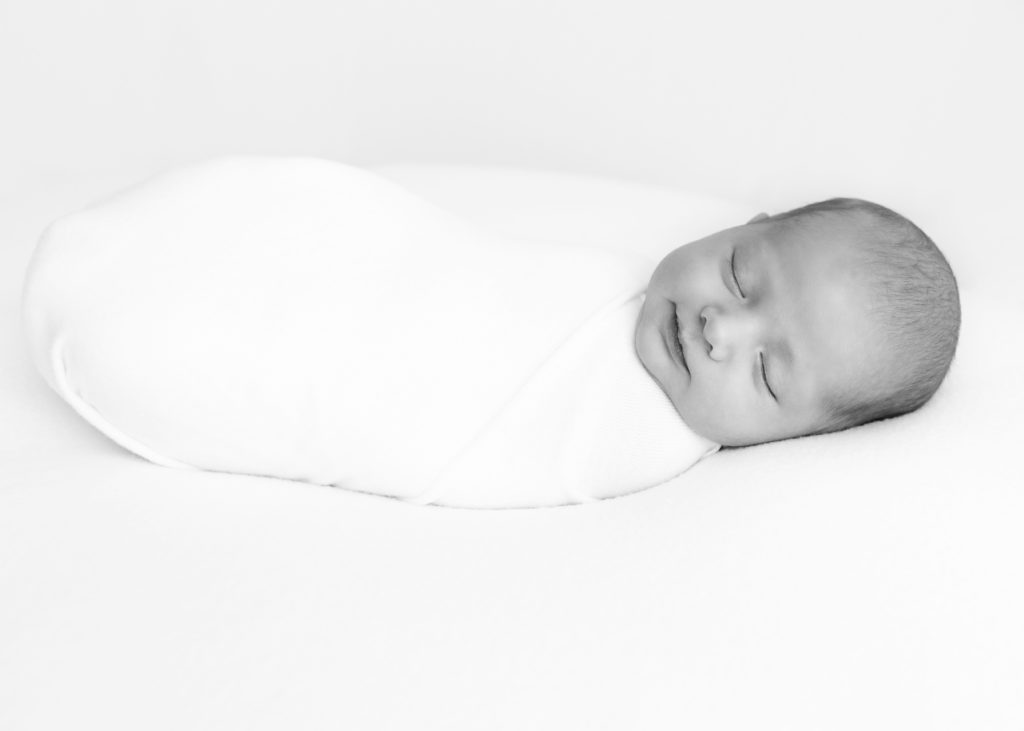Newborn baby boy sleeping and smiling wrapped in white cloth by Grapevine Newborn photographer