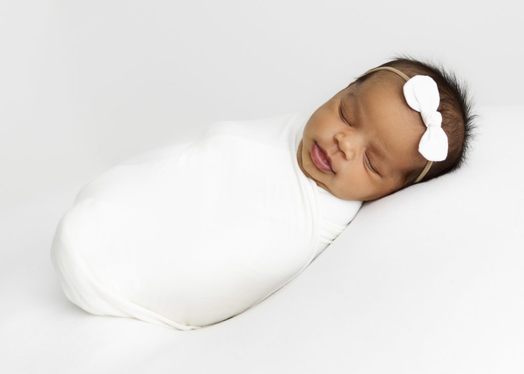 Newborn baby girl wrapped in a white blanket and sleeping with a white bow on her head.