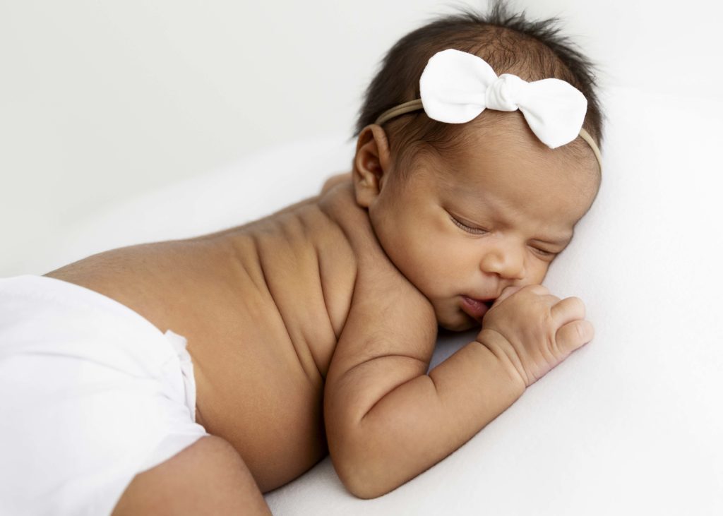 Newborn baby girl sleeping on a white blanket with a white diaper cover on and a simple white bow in her hair
