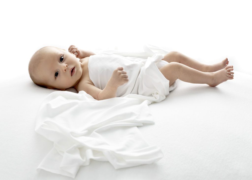 Newborn baby draped in a white blanket looking at the camera