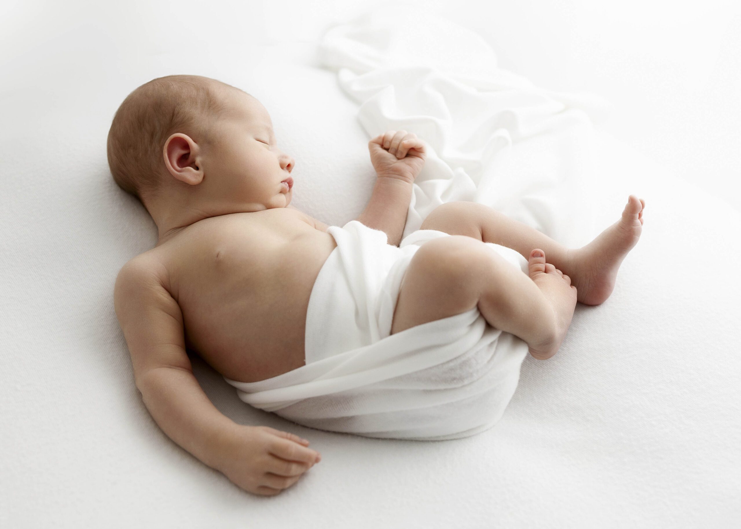 Newborn baby sleeping with white sheet loosely wrapped around him.