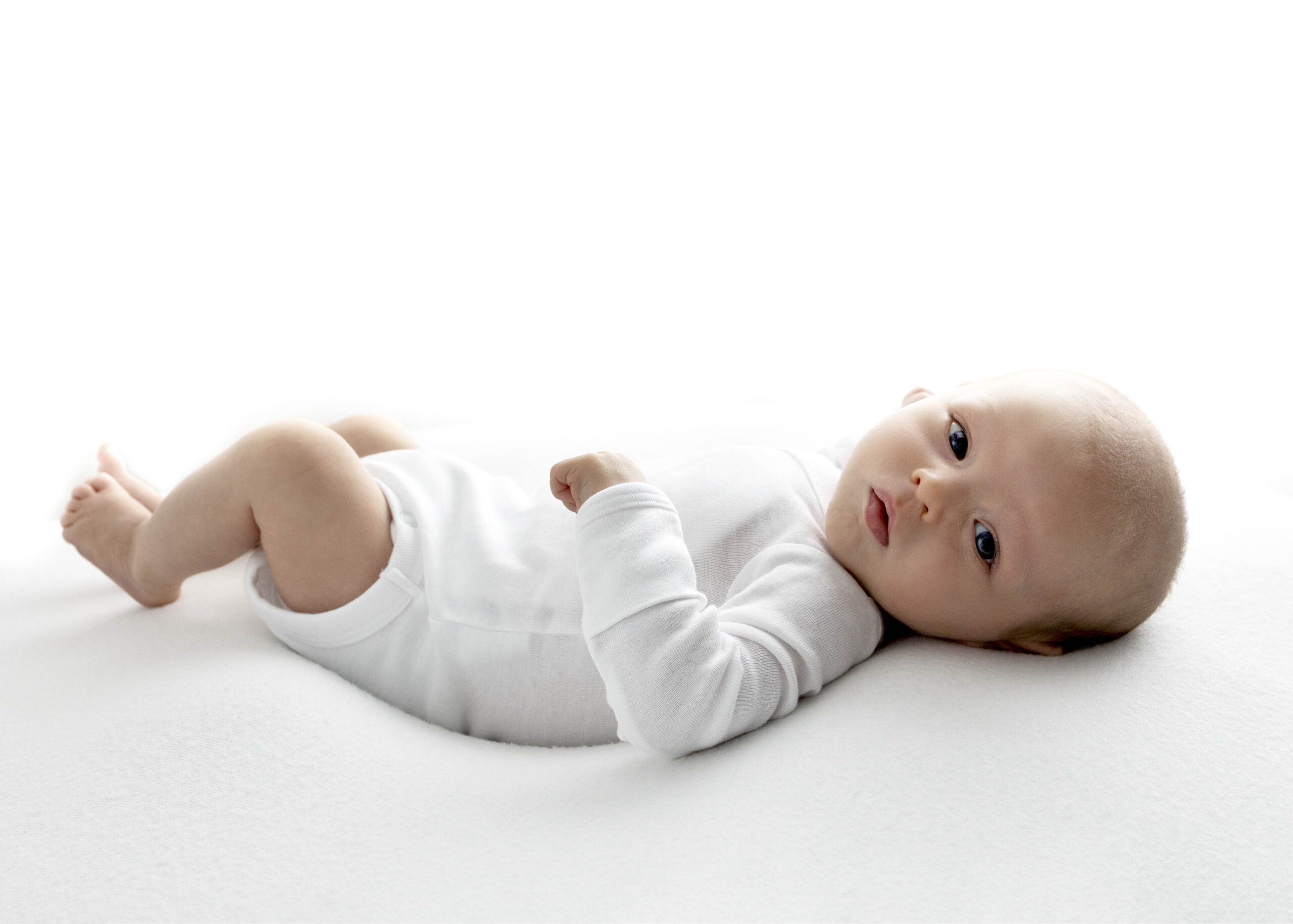 Newborn boy in white onesie lying on back looking at the camera