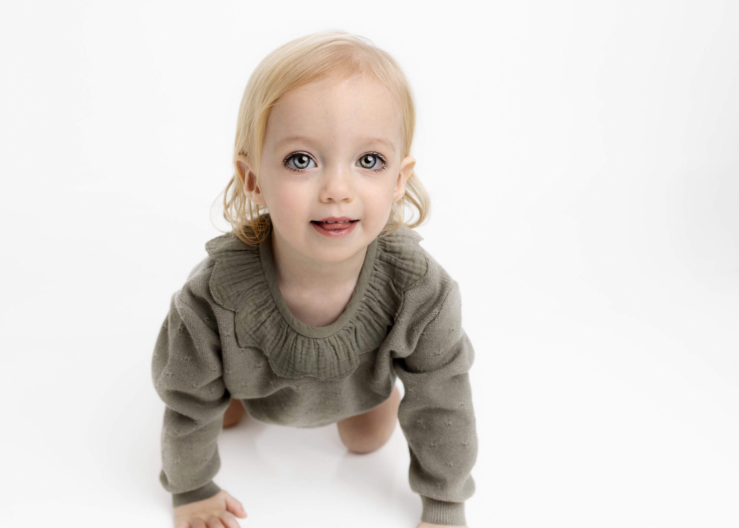 Little girl in green romper on hands and knees looking at the camera by white studio photography
