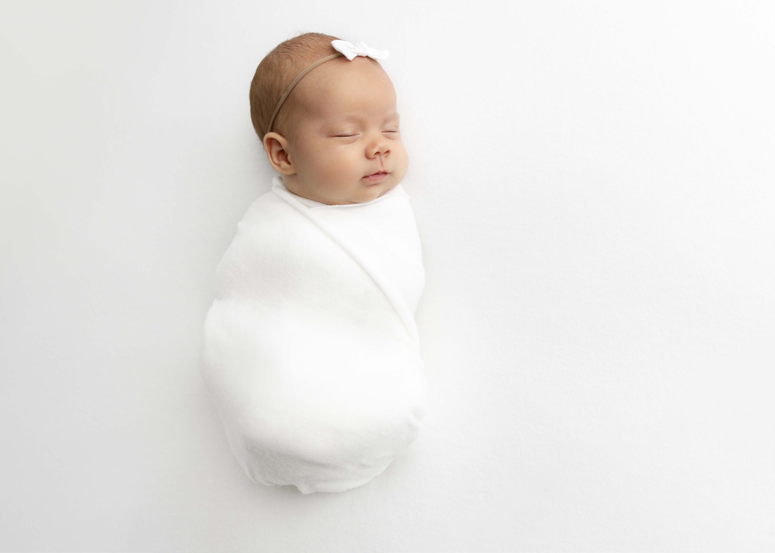 Newborn baby wrapped in white wrap with white bow on her head