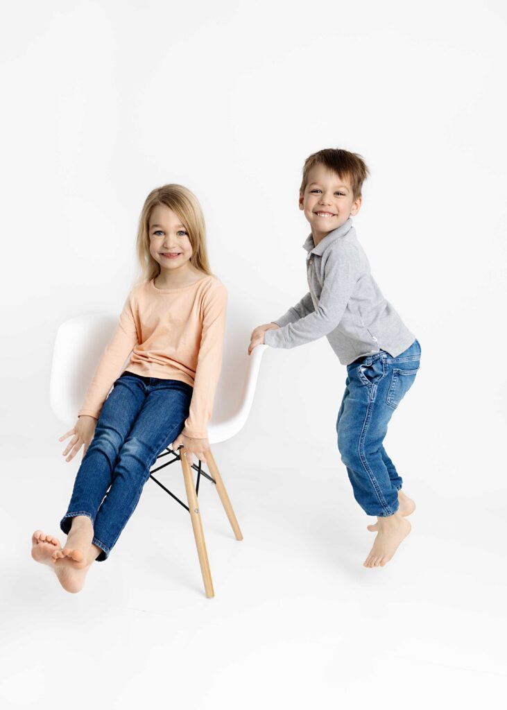 Brother and sister- sister sitting on a chair and brother jumping up while holding on to chair