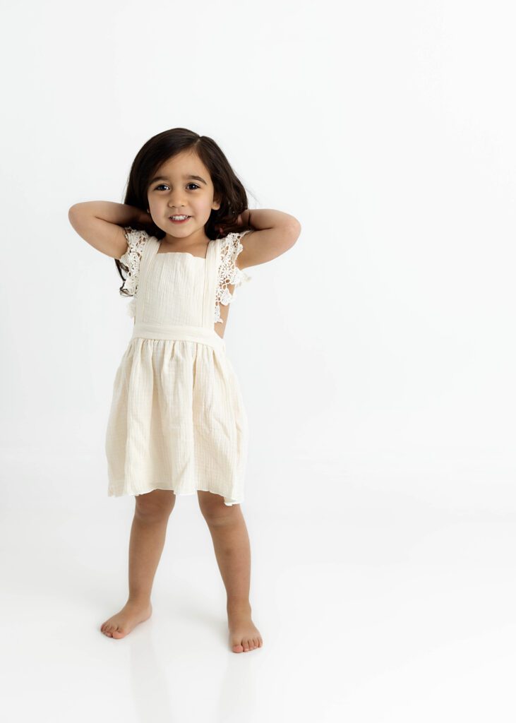 Toddler girl in cream dress standing with arms behind her head