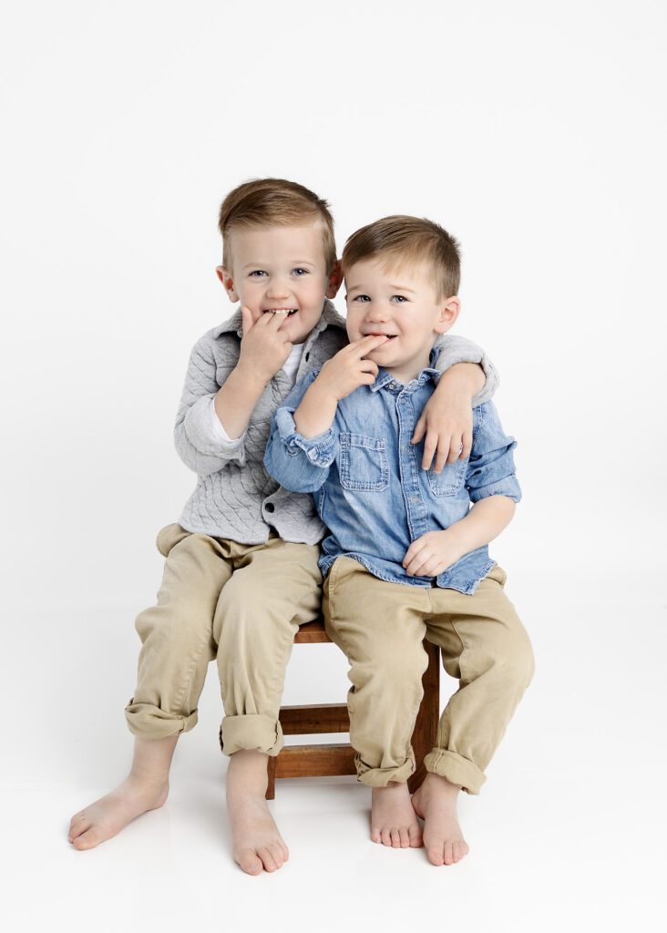 Toddler brothers sitting on stool next to each other with their fingers in their mouths.