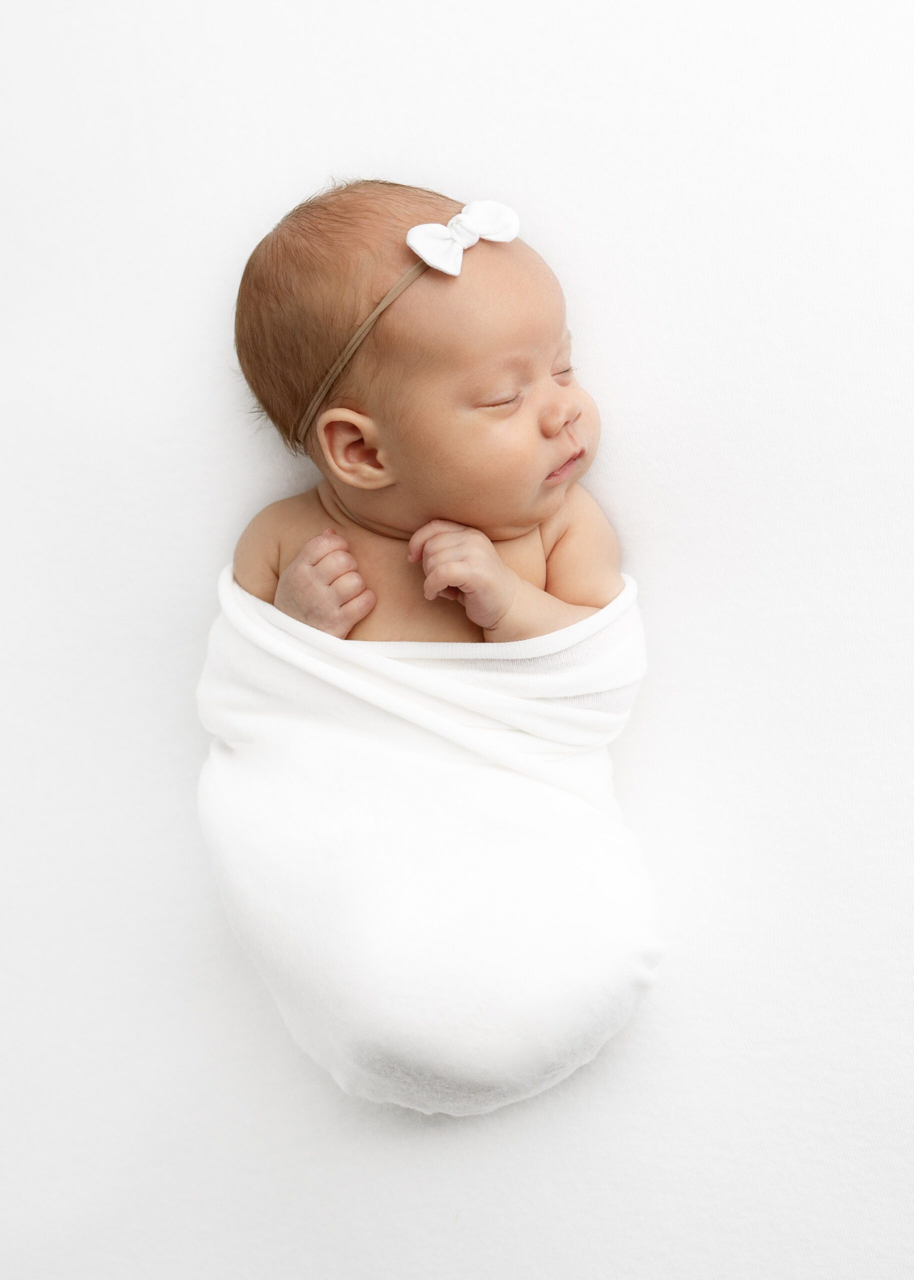 newborn girl swaddled in white blanket with white bow in hair