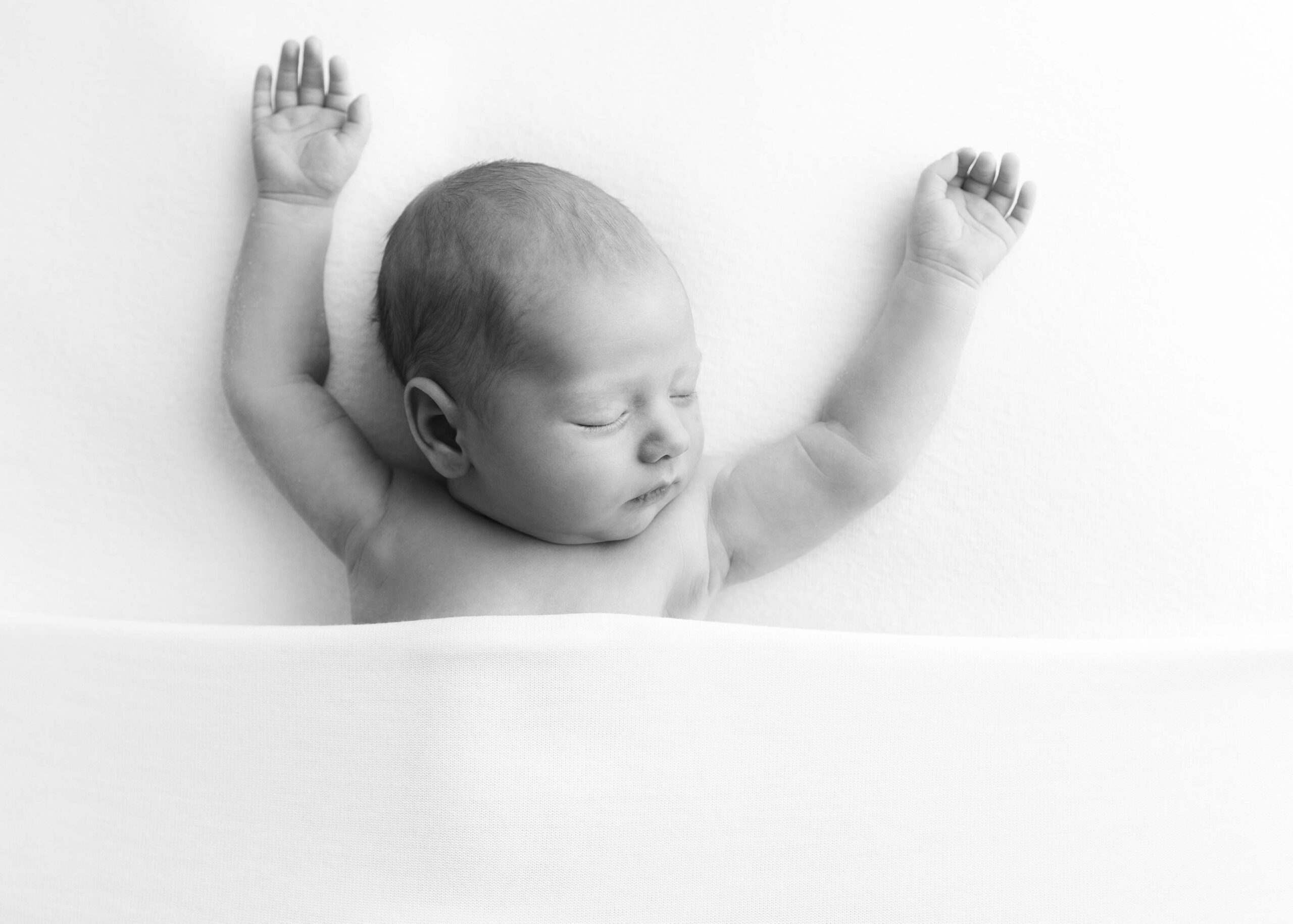 Newborn sleeping under white blanket with arms outstretched