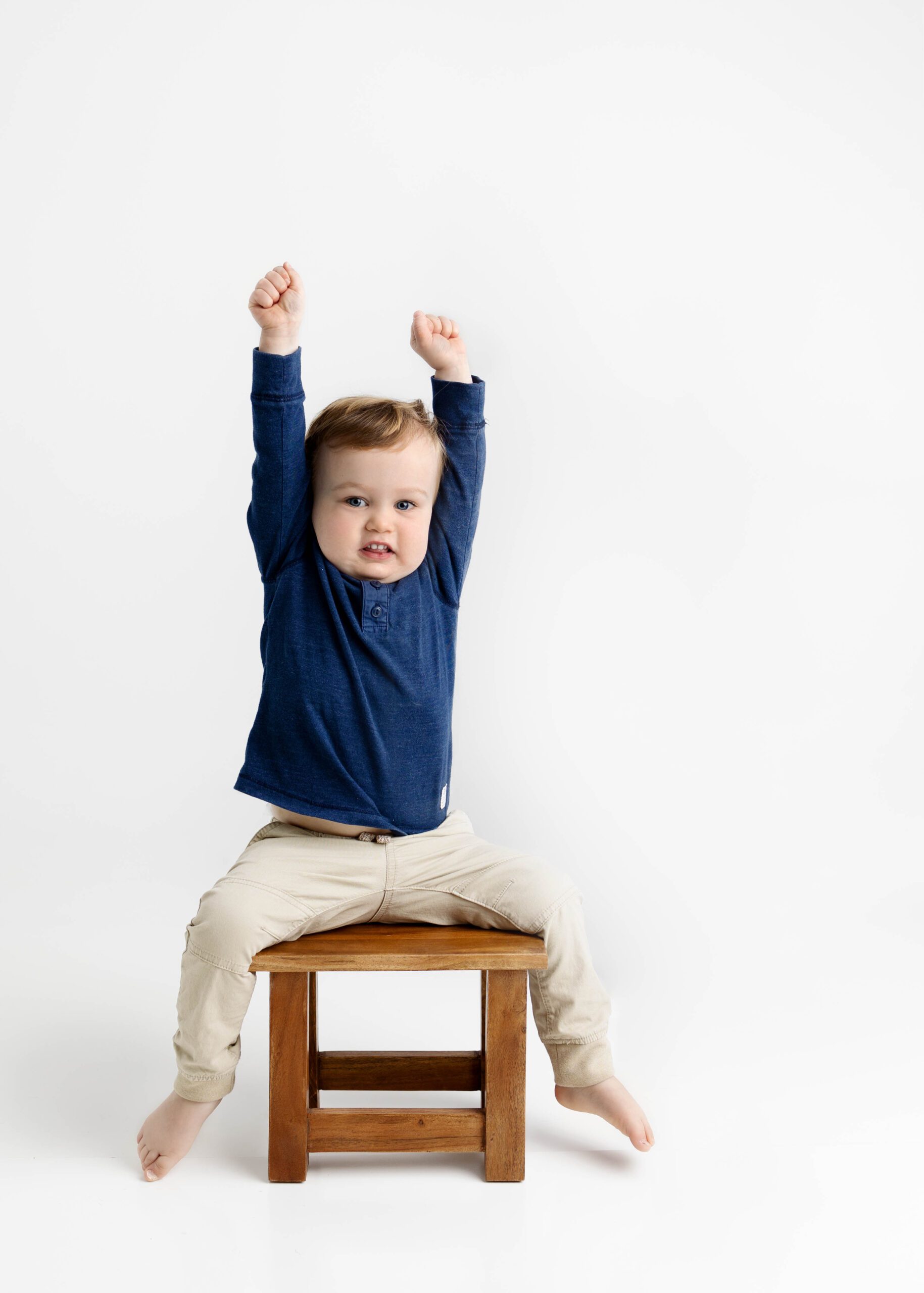 little boy in blue shirt sitting on wood stool with arms stretched above his head