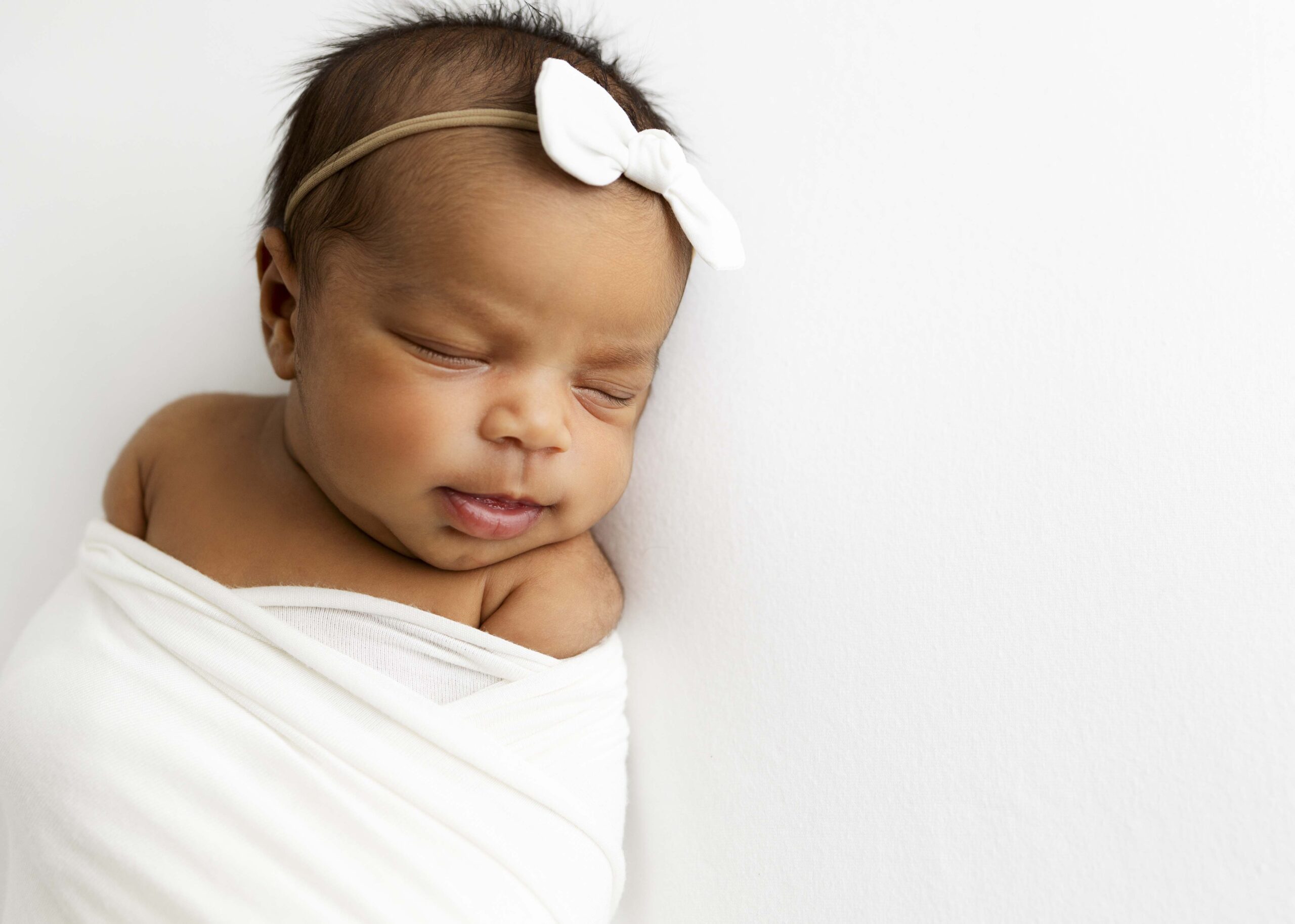 newborn girl with bow in hair and wrapped in a white blanket sleeping